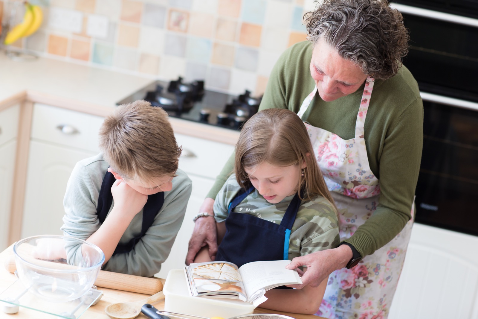 two children cooking with adult in kitchen looking at recipe book