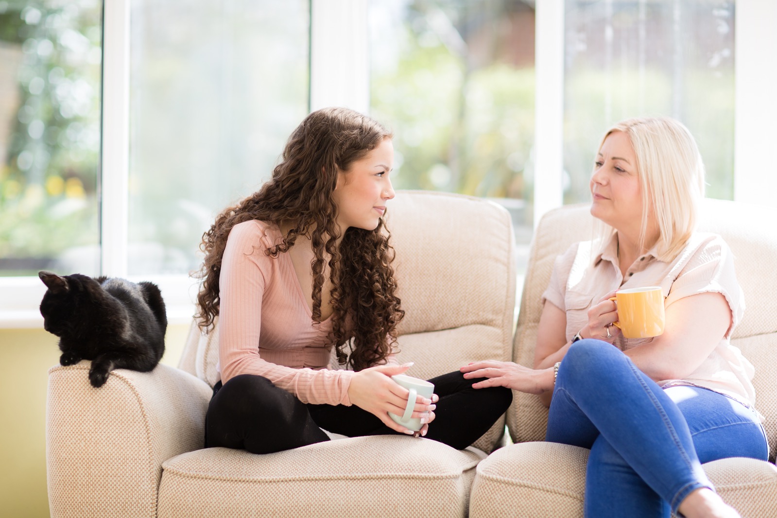 Adult and older teen sitting on sofa having hot drink, with cat on arm rest