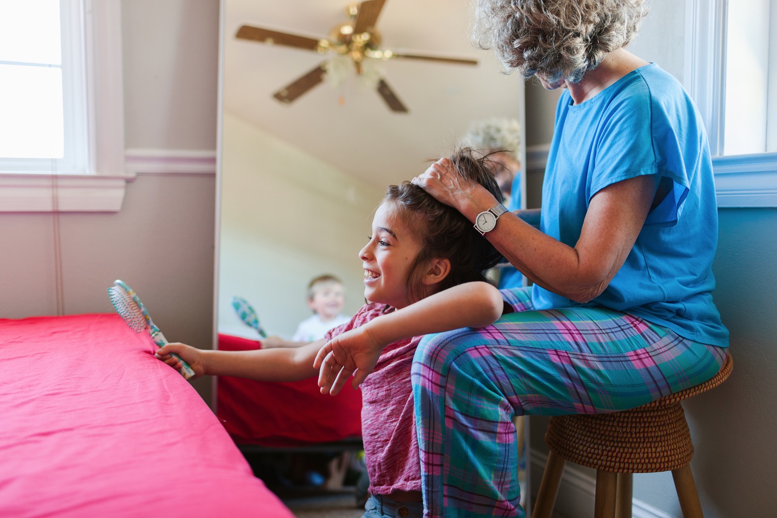 Older lady combing hair of younger girl, younger child looks on in mirror