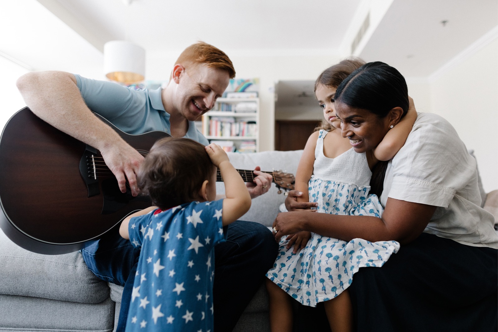 Baby and primary age girl sit on sofa whilst adult male plays guitar